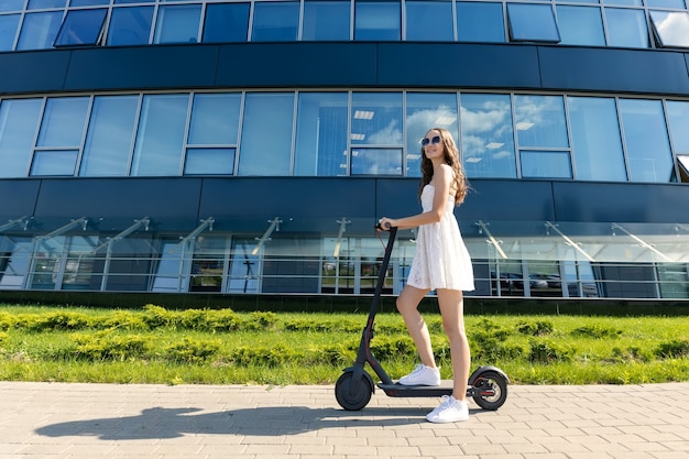 Een meisje in een jurk rijdt op een elektrische scooter in de buurt van moderne gebouwen