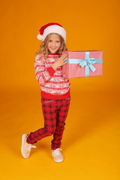 een meisje in een kersttrui in een geruite broek en een kerstmuts houdt een geschenkdoos vast
