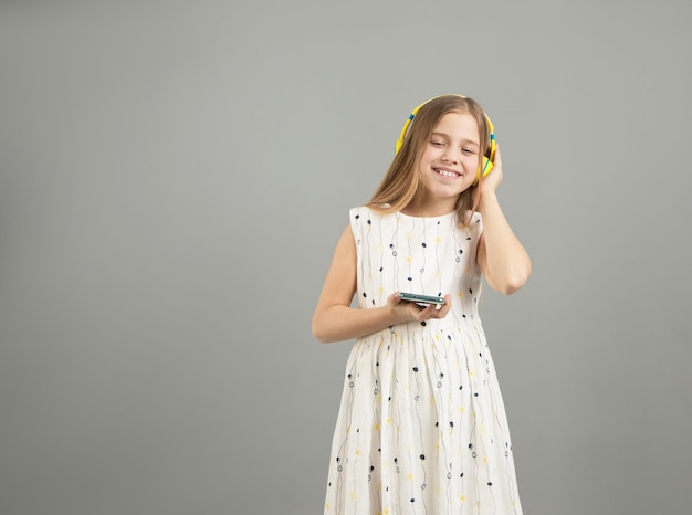 Een meisje in een lichte jurk luistert naar muziek met een gele koptelefoon op een grijze geïsoleerde achtergrond
