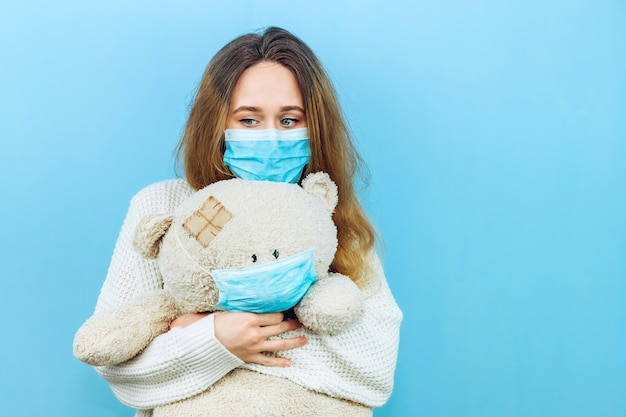 Een meisje in een medisch masker houdt een stuk speelgoed gekleed in een medisch masker op blauw