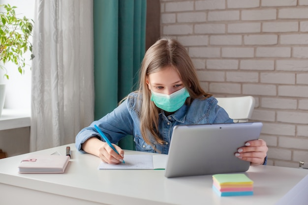 Een meisje in een medisch masker studeert thuis met een digitale tabletlaptop en doet huiswerk. Afstandsonderwijs online onderwijs