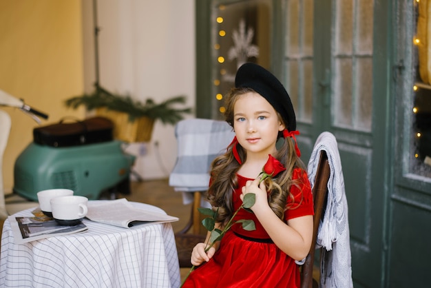 Een meisje in een rode fluwelen jurk zit aan een cafétafel en houdt een rode roos in haar handen