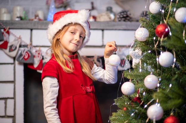 Een meisje in een rode jurk en een kerstman hoed bij de open haard klampt zich vast aan de kerstboom