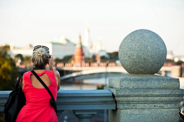 Een meisje in een rode jurk tegen de stenen balustrade staat met haar rug naar ons toe op de achtergrond van het stadslandschap