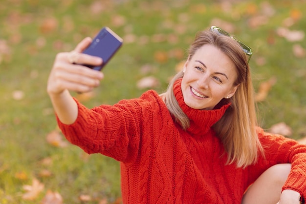 een meisje in een rode trui maakt een selfie op een mobiele telefoon in het herfstpark, het concept van het herfstseizoen