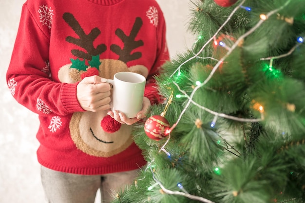 Een meisje in een rode trui met een witte beker in haar handen staat naast de kerstboom in het nieuwe jaar