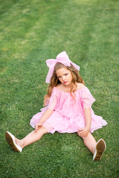 Een meisje in een roze jurk met een grote strik op haar hoofd zit op het gras. Meisje pop