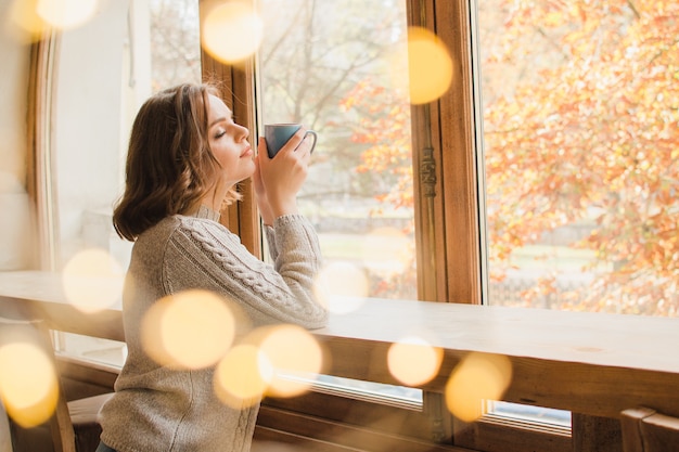 Een meisje in een trui drinkt thee of koffie en spleet uit het raam