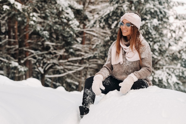 Een meisje in een trui en bril in de winter zit op een besneeuwde achtergrond in het bos.