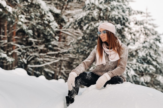 Een meisje in een trui en een bril in de winter zit op een besneeuwde achtergrond in het bos