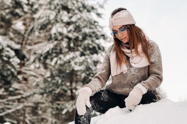 Een meisje in een trui en een bril in de winter zit op een besneeuwde achtergrond in het bos