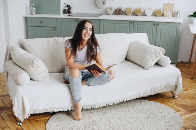 Een meisje in een wit T-shirt en een gescheurde spijkerbroek zit op de bank met een boek in haar hand en lacht breed