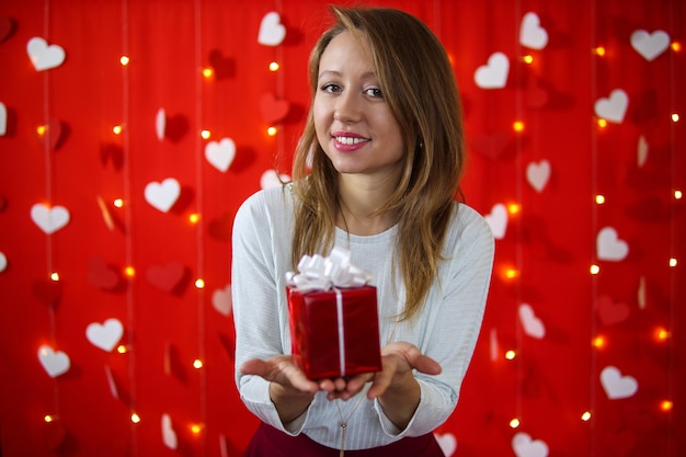 Een meisje in een witte blouse en rok kijkt naar een cadeau in haar hand op een rode achtergrond met hartjes. Valentijnsdag concept