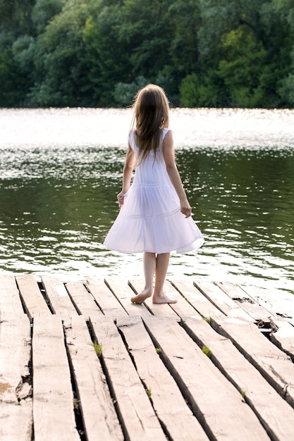 Een meisje in een witte jurk staat op een houten pier bij de rivier. Uitzicht vanaf de achterkant