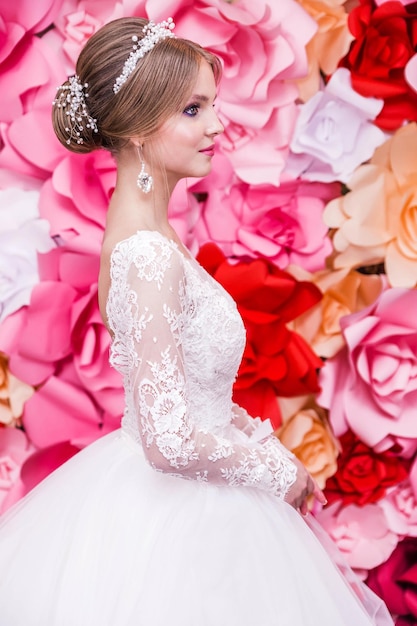 Een meisje in een witte trouwjurk met een open rug make-up een kapsel in de vorm van een knot poseert op een felrode achtergrond van papieren bloemen