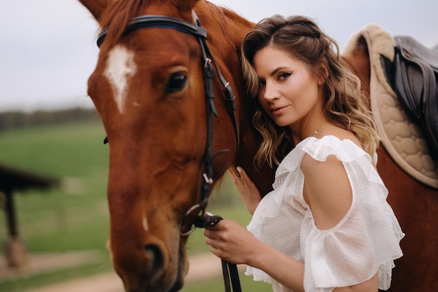 Een meisje in een witte zomerjurk staat in de zomer naast een bruin paard in een veld