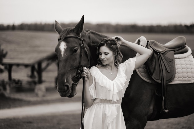 Een meisje in een witte zomerjurk staat naast een bruin paard in een veld in de zomer zwart-wit foto