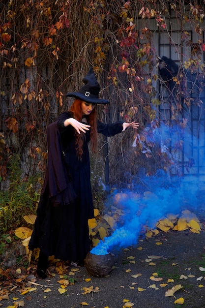 een meisje in een zwart heksenkostuum en een puntmuts met een pot blauwe rook op Halloween in de herfstpark