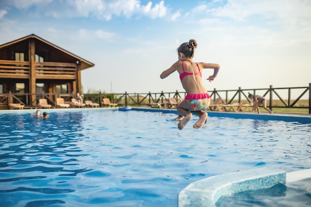 Een meisje in felroze zwembroek en zwarte zwembril, grappig springend in diepblauw zwembad met schoon transparant koel water tegen de achtergrond van de warme zomerzonsondergang