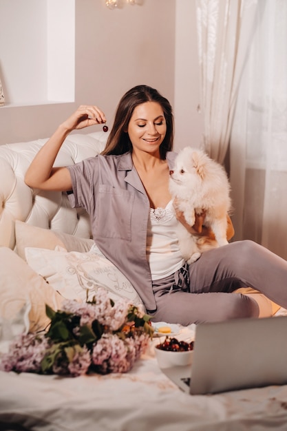 Een meisje in pyjama's zit 's nachts in bed met haar witte hond die naar een laptop kijkt en snoep eet. Meisje met een hond Spitzer thuis in bed.