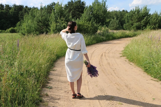 Een meisje in witte kleren loopt langs een landweg met een boeket wilde bloemen