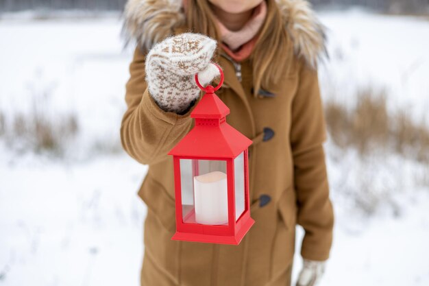 Een meisje in wollen wanten houdt een rode lantaarn in haar handen met een kaars in het winterbos
