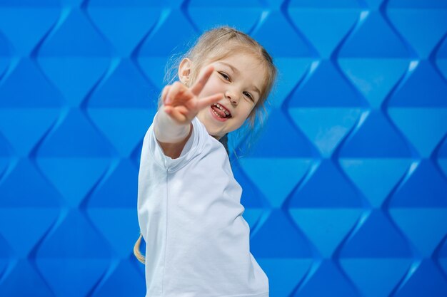 Een meisje met blond haar in een wit T-shirt op een blauwe achtergrond. Ze lacht en zwaait met haar handen