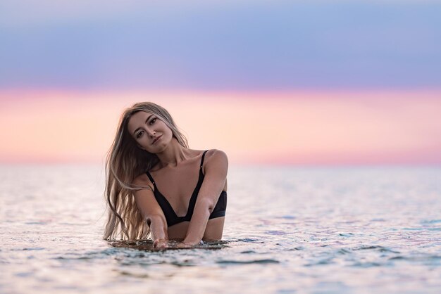 Een meisje met blond haar in een zwart zwempak spettert naar de zijkanten terwijl ze in een estuarium zit op de achtergrond van een zonsondergang