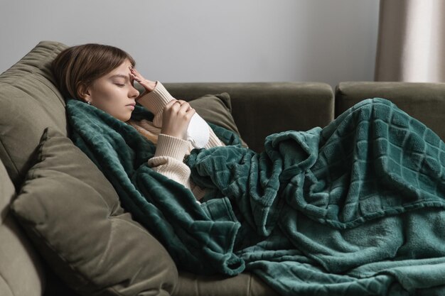 Een meisje met de eerste symptomen van de ziekte ligt bedekt met een deken op de bank en houdt haar voorhoofd vast, de temperatuur is gestegen