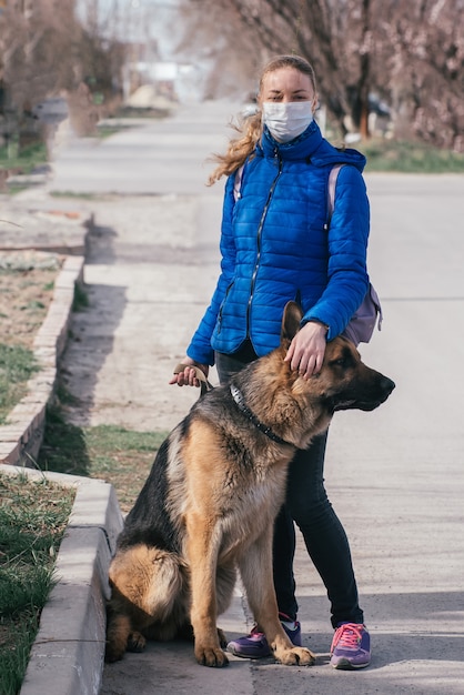 Foto een meisje met een beschermend medisch masker laat een hond op straat lopen. vrije tijd met een huisdier tijdens quarantaine. loop met een duitse herder