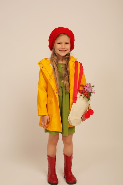 een meisje met een gele regenjas rode laarzen een groene jurk een rode baret houdt een portemonnee met wilde bloemen vast