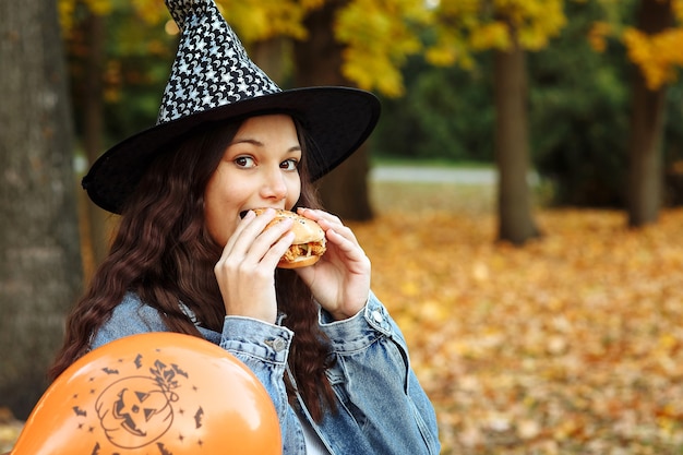 een meisje met een heksenhoed naast een oranje ballon voor Halloween eet een hamburger in een herfstpark