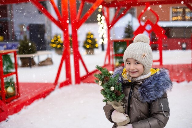 Een meisje met een kerstboom in haar handen buiten in warme kleren in de winter op een feestelijke markt Sprookjesslingers versierde sneeuwstad voor het nieuwe jaar