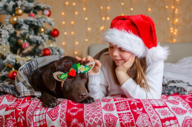 Een meisje met een kerstman-hoed met een hond ligt op het bed bij de kerstboom