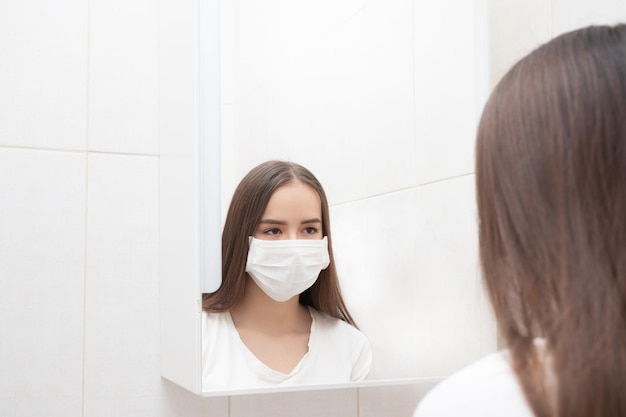 Een meisje met een medisch masker kijkt thuis in de spiegel in de badkamer Preventie en bescherming tegen ziekten