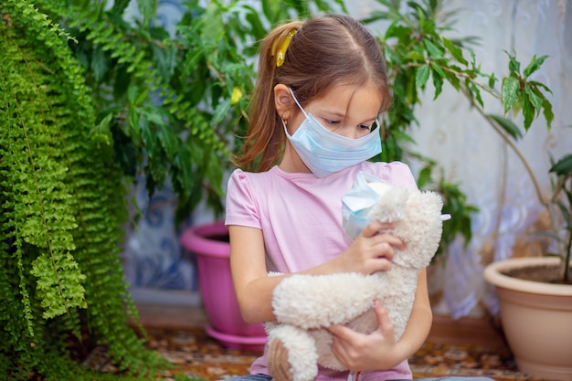 Een meisje met een medisch masker speelt thuis met haar teddybeer, die ook in een masker zit