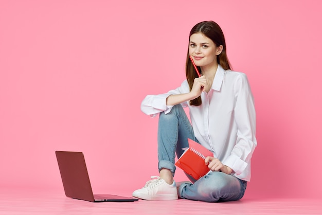 Een meisje met een mooie glimlach zit op een roze achtergrond met een laptop en kijkt naar de camera