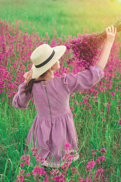 Een meisje met een strohoed en een paarse jurk zit in een veld en geniet van de geur van een boeket paarse bloemen