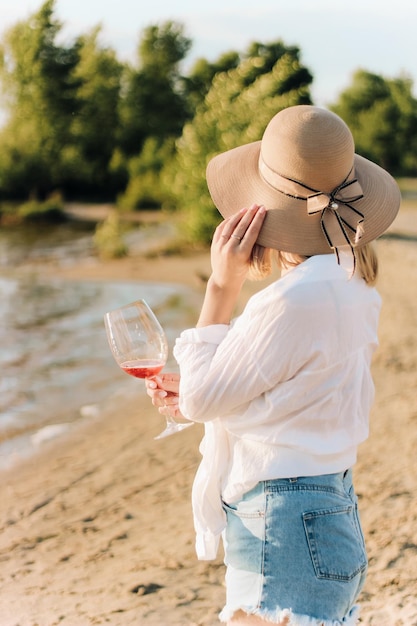 Een meisje met een strohoed met een glas wijn zit op het strand bij zonsondergang