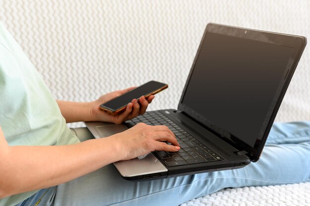 Een meisje met een telefoon in haar handen is bezig thuis op een laptop met een kopje op de bank met licht uit het raam