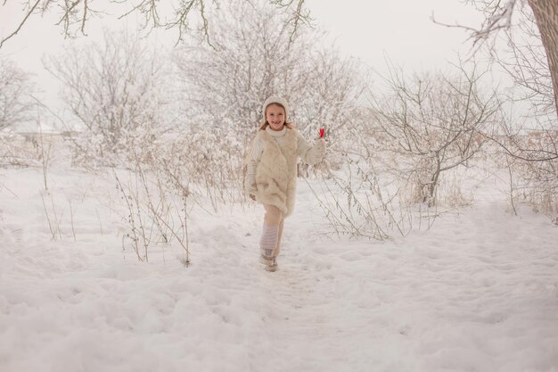 een meisje met een witte gebreide muts en trui met een snoepje in haar handen in een besneeuwd park