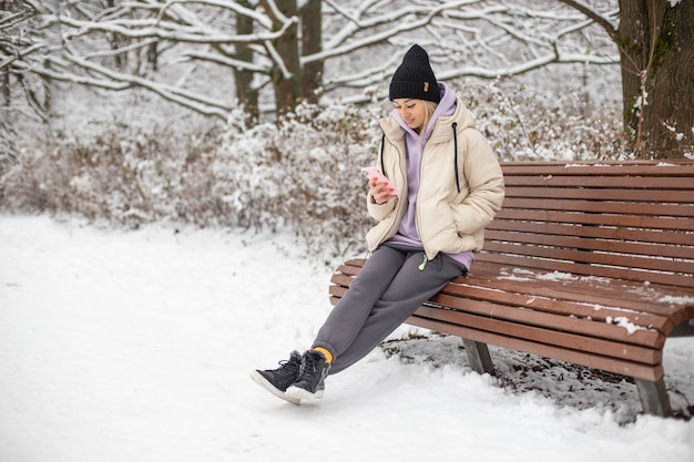 Een meisje met een zwarte hoed zit op een bankje in het winterpark en checkt berichten op haar telefoon