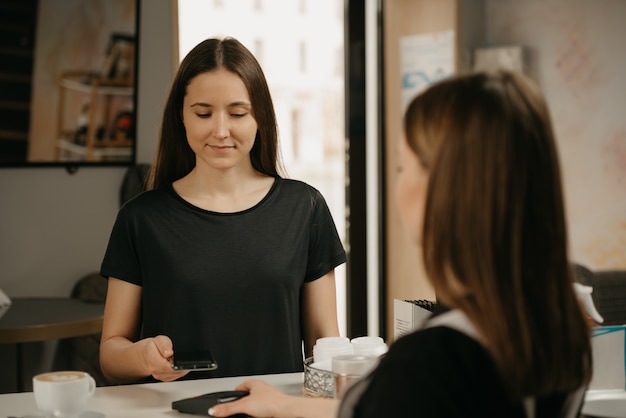 Een meisje met lang haar betaalt voor haar koffie met een smartphone door contactloze NFC-technologie in een café. Een vrouwelijke barista reikt een cliënt een betaalterminal aan.