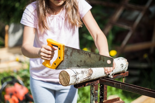 Een meisje met lang haar helpt ouders in de zomer, zaagt een houtblok met een zaag, hout, constructie, brandhout, kachel, spel, training