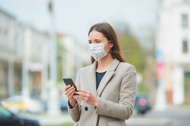 Een meisje met lang haar in een medisch gezichtsmasker om de verspreiding van het coronavirus te voorkomen, gebruikt een smartphone op straat. Een vrouw in een gezichtsmasker tegen COVID-19 draagt een jas in het centrum van de stad.