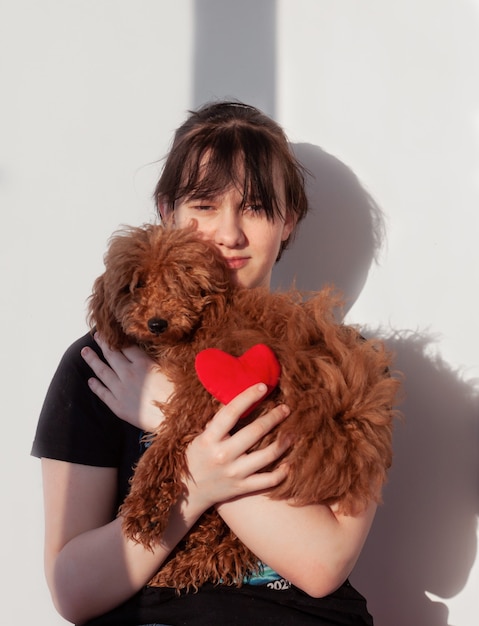 Een meisje met zwart haar knuffelt een ruige roodbruine poedelhond en houdt een rood hart in haar hand.