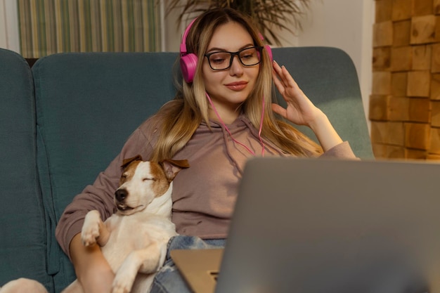 Een meisje op de bank ligt met een hond in haar laptop en luistert naar muziek via een koptelefoon Freelancen en communiceren via internet