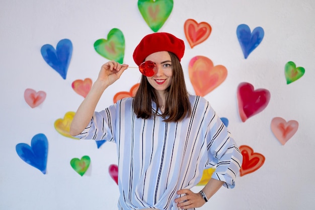 Een meisje op een achtergrond van kleurrijke heldere harten met een rood lollyhart glimlacht terwijl ze haar oog sluit het concept van valentijnsdagliefde