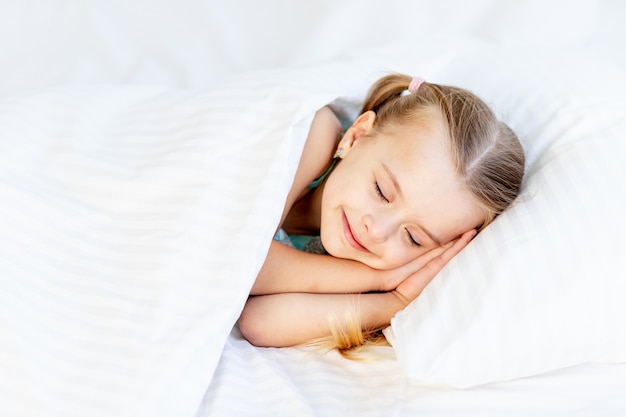 Een meisje slaapt thuis op een bed op een wit katoenen bed en lacht liefjes in haar slaap met haar handen gevouwen onder haar wangen