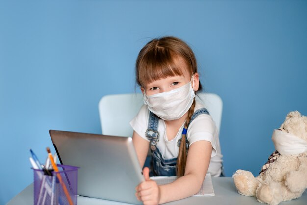 Een meisje van 5-6 jaar dat aan een tafel zit, voert huiswerk afgelegen taken uit op de tablet. Isoleer op een blauwe muur. Coronavirus, gemaskerd kind.